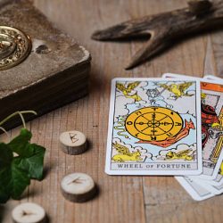 A moody, atmospheric scene featuring tarot cards laid out on an old wooden table, with the "Wheel of Fortune" card placed on top. Surrounded by rune stones and an ancient book embossed with a Celtic triquetra symbol. Warm lighting and dark, mystical tones create a spiritual and magical ambiance. Perfect for themes like divination, mysticism, witchcraft, or the occult.